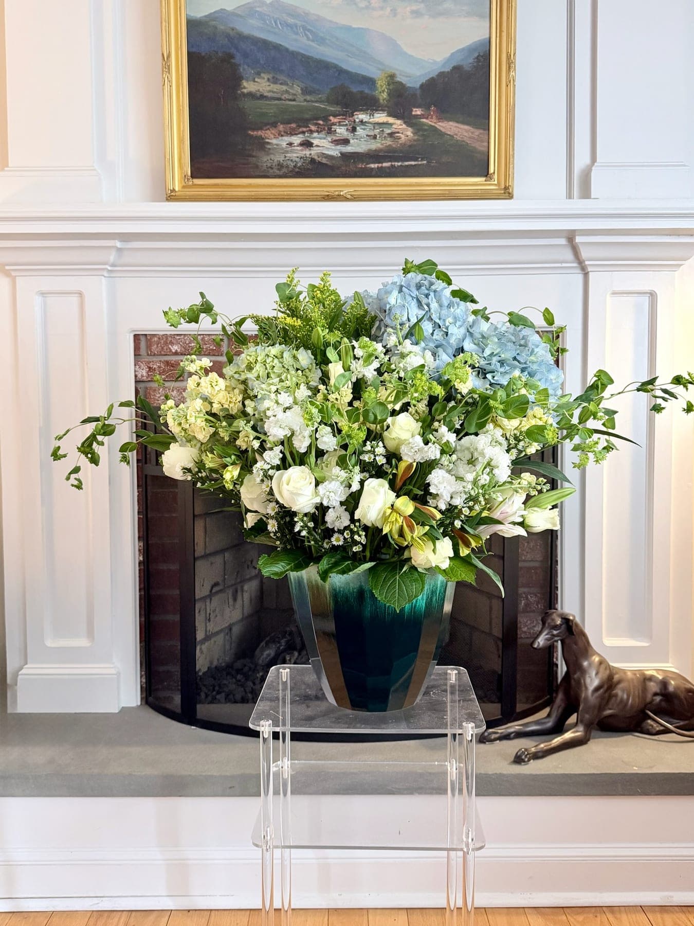 Large arrangement of blue hydrangea, white stock, and roses in a teal glass vase on a mantel