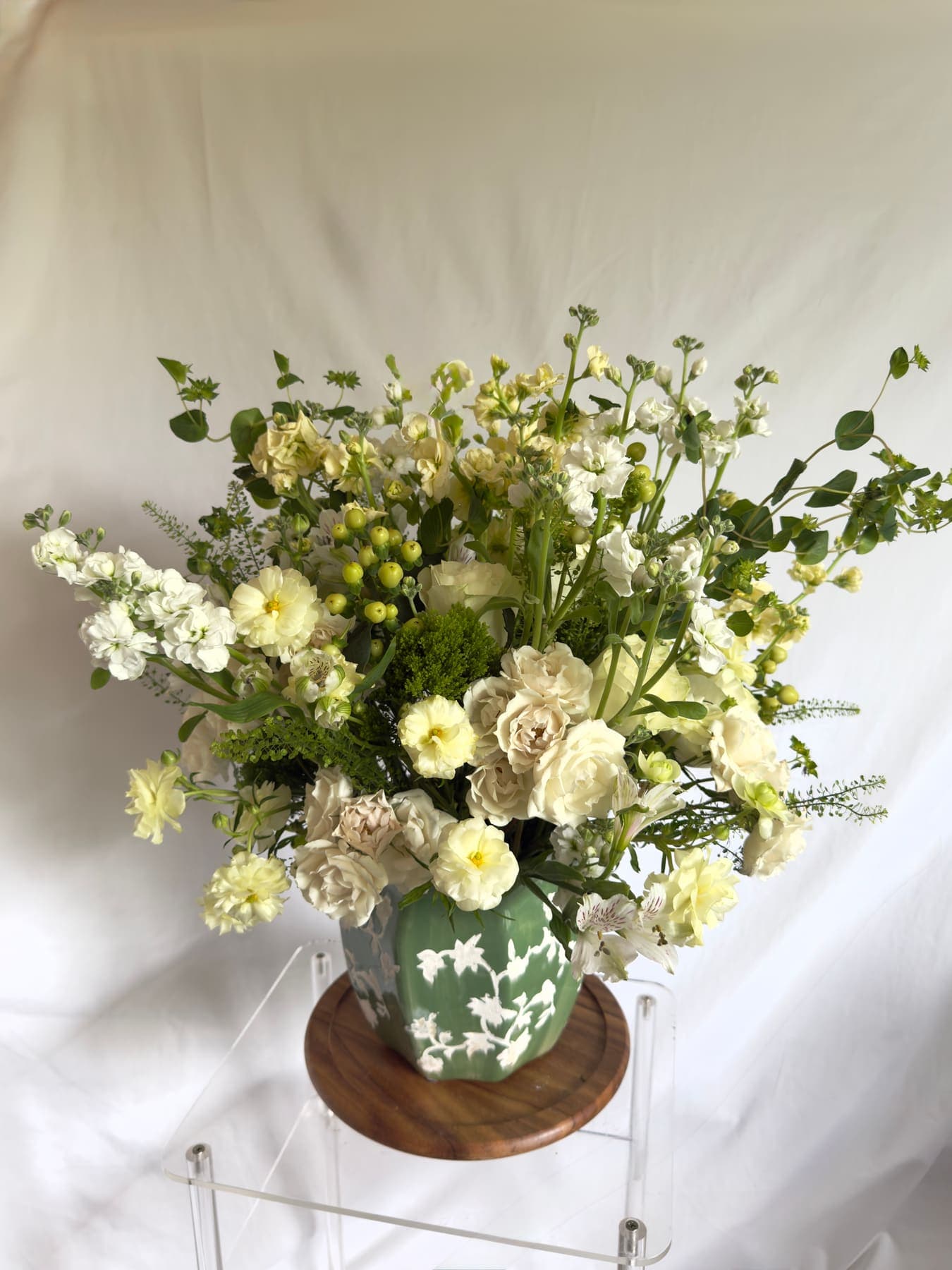 Cream and green arrangement spilling over a celadon vase on a wooden stand