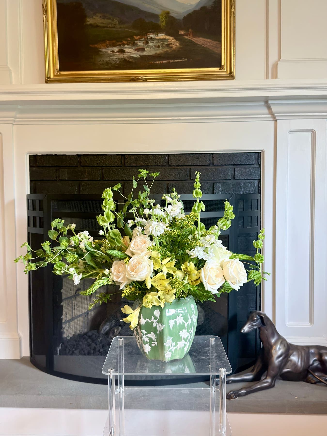 Arrangement in a green vase in front of a fireplace beneath a gilt-framed painting