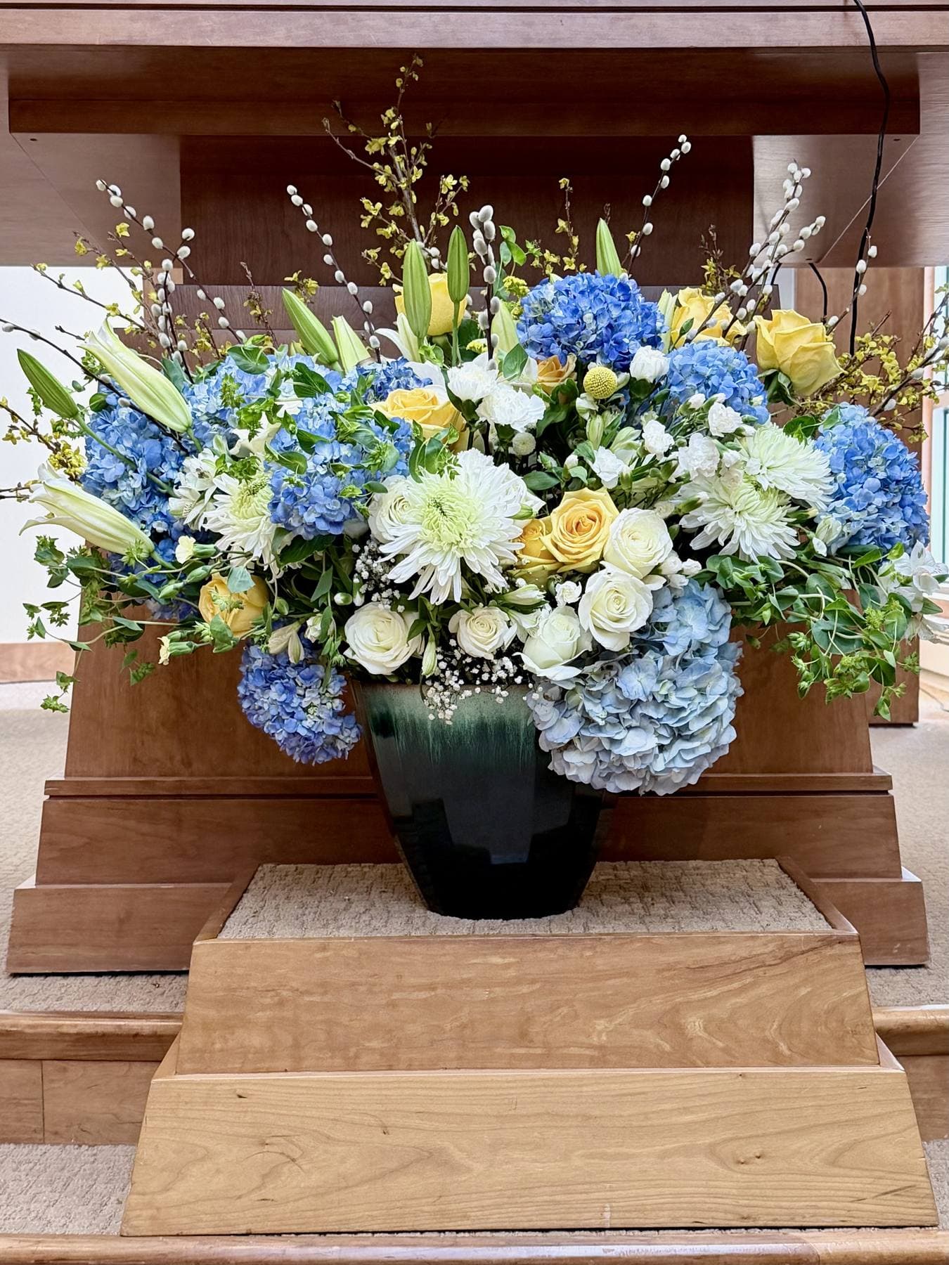 Large arrangement of blue hydrangeas, white and yellow roses, and pussy willow in a green urn